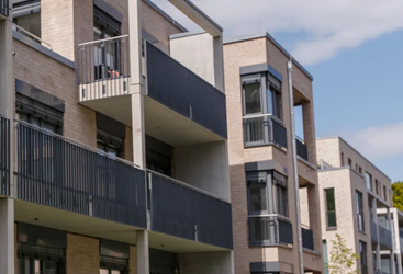 Moderne Wohnhäuser mit mehreren Stockwerken, Balkonen und großen Fenstern. Die Fassaden bestehen aus hellem Klinker und dunklen Metallelementen. Die Architektur wirkt zeitgemäß und urban.

                                  